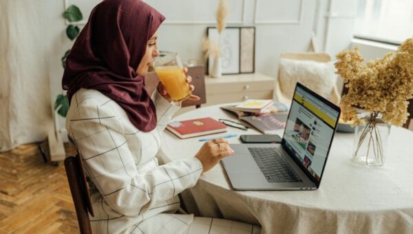 wanita berhijab bekerja dari rumah sambil minum jus di dekat laptop, ilustrasi kerja dari rumah untuk ibu rumah tangga